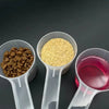 Three transparent measuring spoons hold dry dog and cat food pellets or powder on a dark surface.