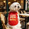 A white dog wearing a red pet sweater with the text I Love my Mommy sitting on a wooden chair in a cafe.