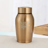 A gold-colored stainless steel pet memorial storage box with a black lid sitting on a light surface.