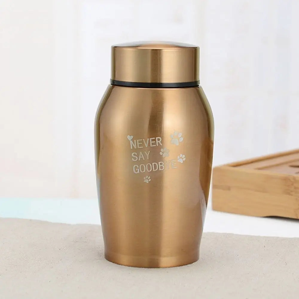 A gold-colored stainless steel pet memorial storage box with a black lid sitting on a light surface.