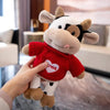 A hand is holding a plush toy of a cow with black and white markings and a red sweater.