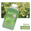 A clear bottle filled with green cat litter deodorizing beads, labeled Apple, set against a blurred outdoor background of gre