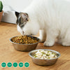 A white cat is eating from a stainless steel pet feeding bowl placed on a wooden surface.