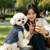 A young woman is holding a white poodle wearing a denim pet jacket outdoors.