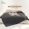 A gray waterproof dog bed cover is shown with a pet resting on it, placed on a light-colored bedding.