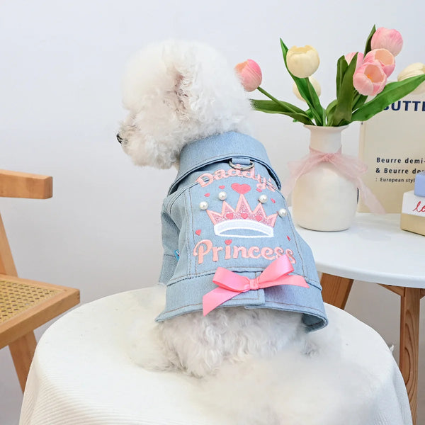 A fluffy white dog wearing a light blue denim jacket decorated with a pink crown and bow, sitting on a white surface.