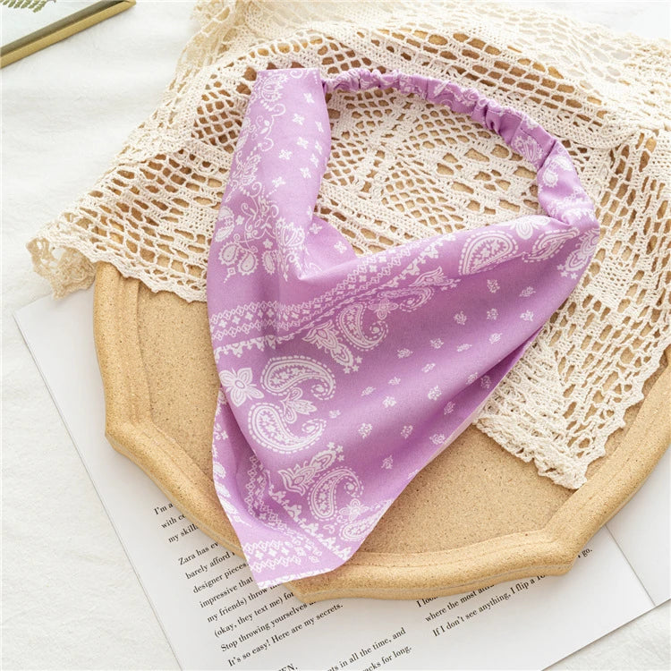 A purple bohemian paisley triangle head scarf is displayed on a light woven fabric and next to some paper.