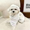 A white dog wearing a white multi-purpose pet bath robe, lying on a light surface.