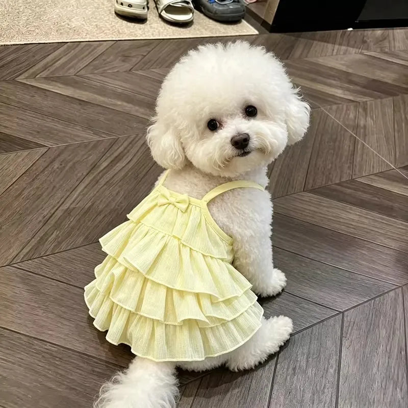 A fluffy white dog wearing a yellow ruffled strap dress sits on a gray tiled floor.