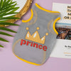 A gray mesh pet vest with a gold crown print and yellow trim resting on a pink surface.