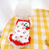 A white pet vest with a red trim and cherry print decoration lying on a yellow checkered surface.