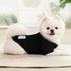 A white dog wearing a black knitted winter sweater, lying down on a light surface.