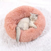 A light gray kitten is resting inside a round, fluffy pink plush pet bed on a white surface.
