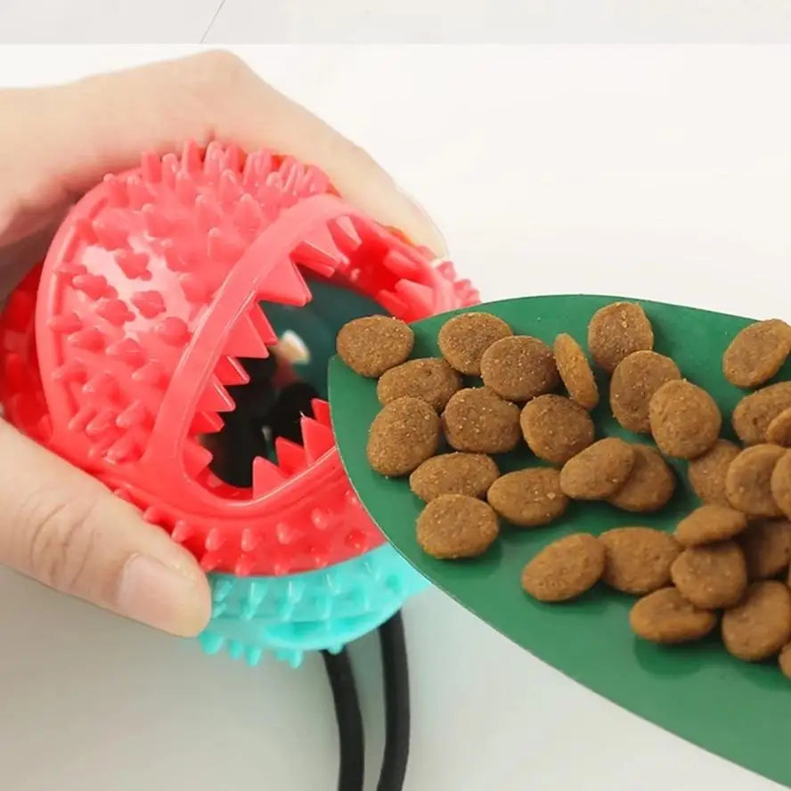 A hand holds a red plastic chew toy with jagged teeth and a blue suction cup base next to a pile of brown dog treats.