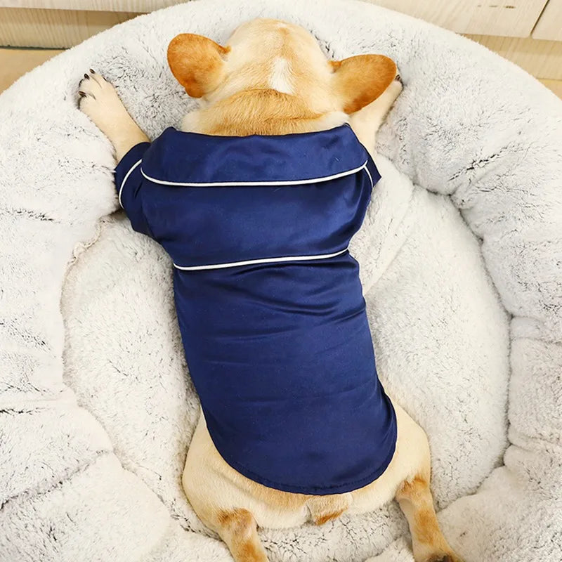 A light-colored dog is lying in a white fluffy bed wearing a navy blue silk pet pajama top.