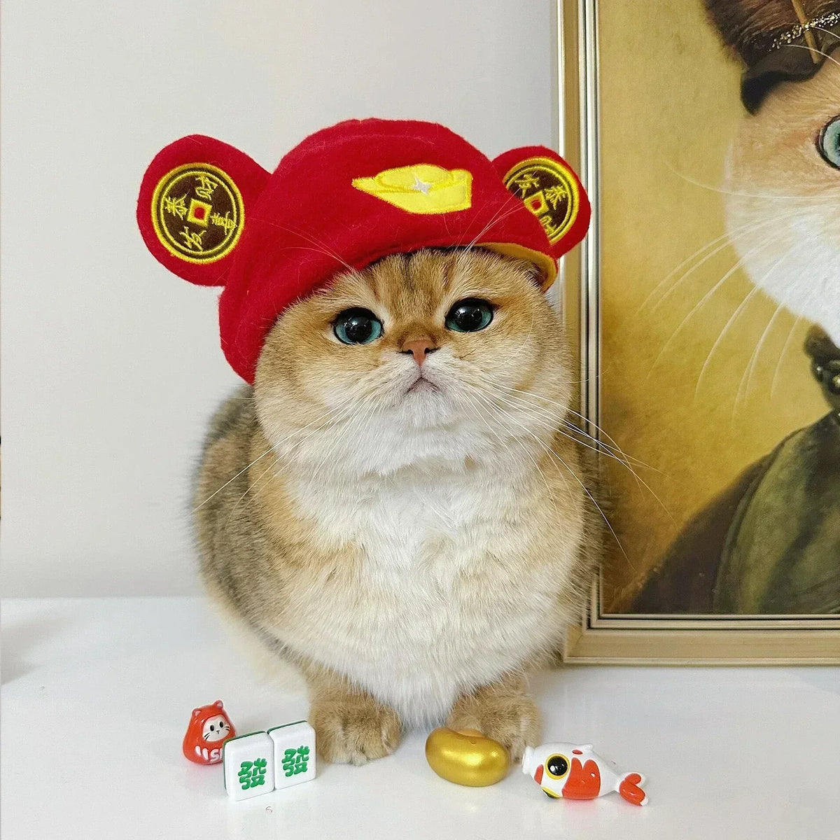 Ein flauschiger, orange Kater mit rojem Hut und goldenen Akzenten sitzt auf einer weißen Oberfläche neben kleinen Spielzeugen