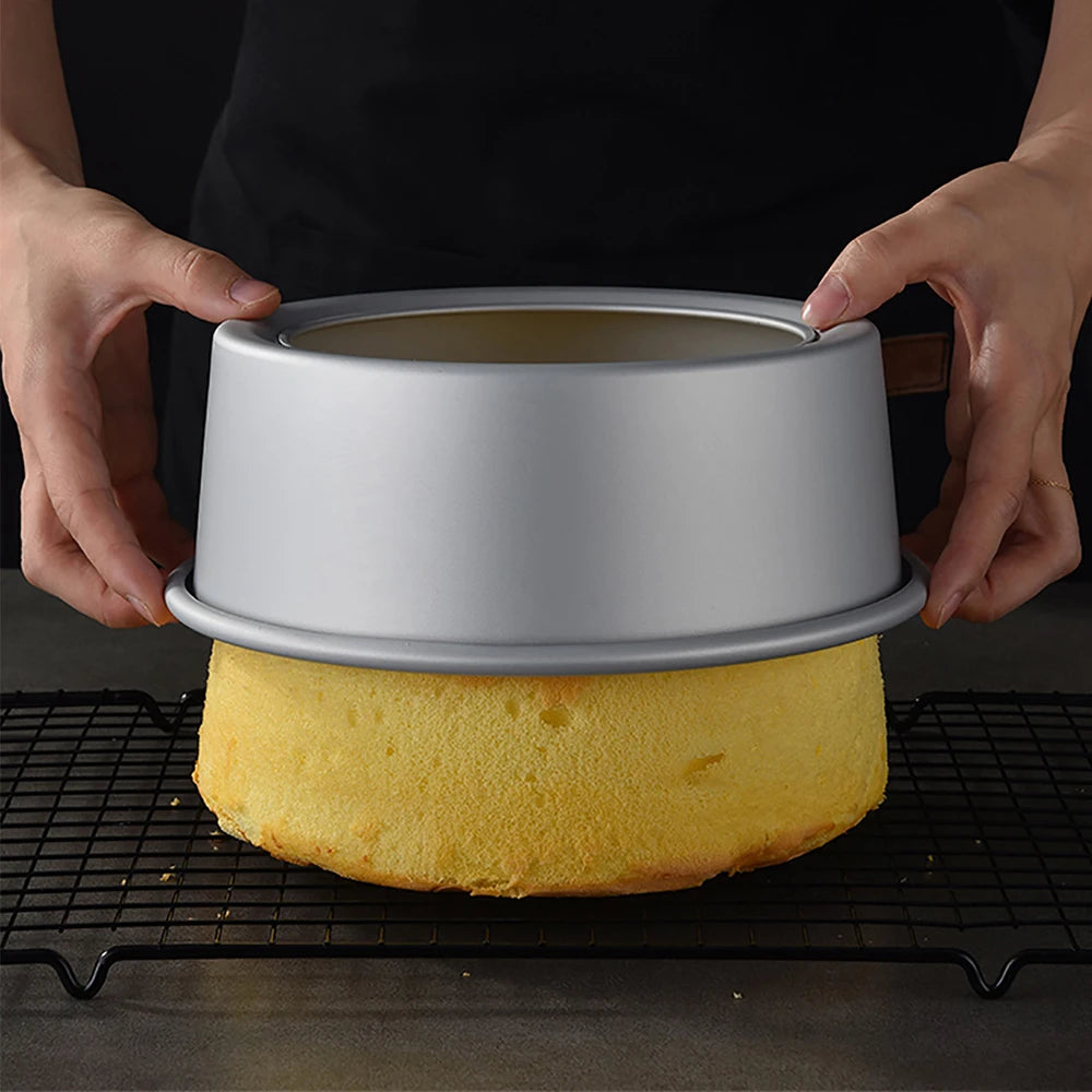 Hands hold a round aluminum cake pan set with a yellow cake inside on a black grill grate.