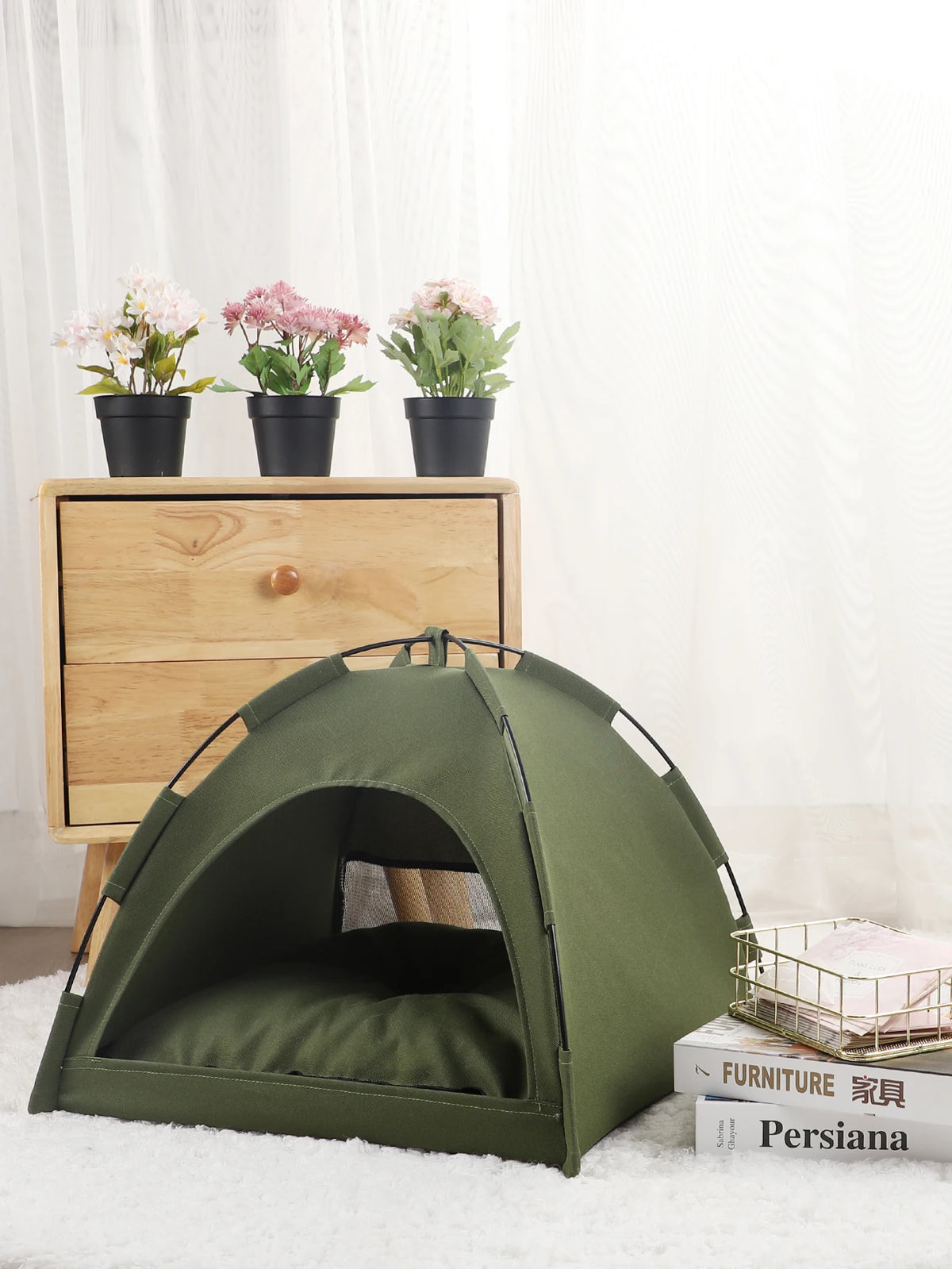 A olive green cozy pet tent bed for cats resting on a white fluffy rug next to potted flowers.