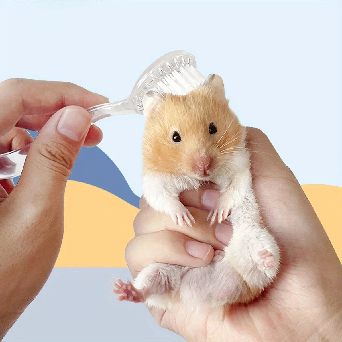 Eine Hand hält eine weiche Bürste für Hamster bei einem kleinen, hellbraunen Hamster.