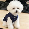 A fluffy white dog wearing a navy blue sleeveless vest with cartoon embroidery, sitting on a wooden desk.
