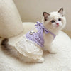 A small white and gray kitten wearing a purple floral print dress with thin straps, sitting on a white fluffy surface.