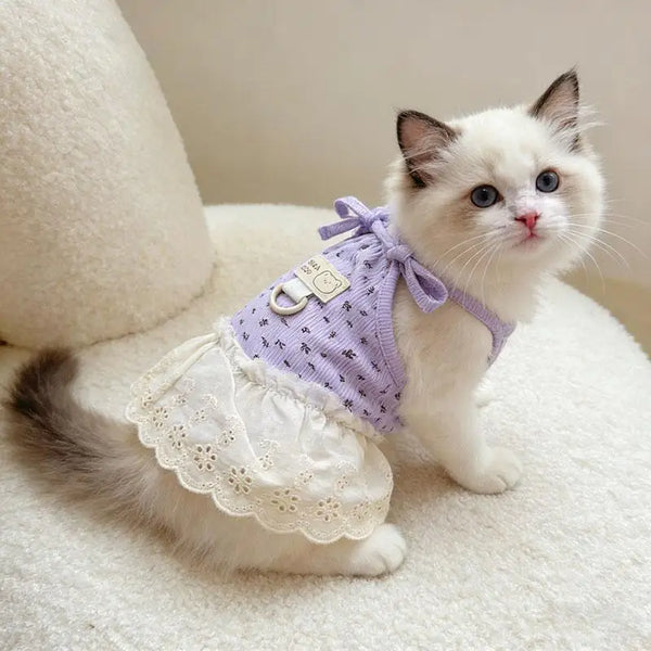 A small white and gray kitten wearing a purple floral print dress with thin straps, sitting on a white fluffy surface.