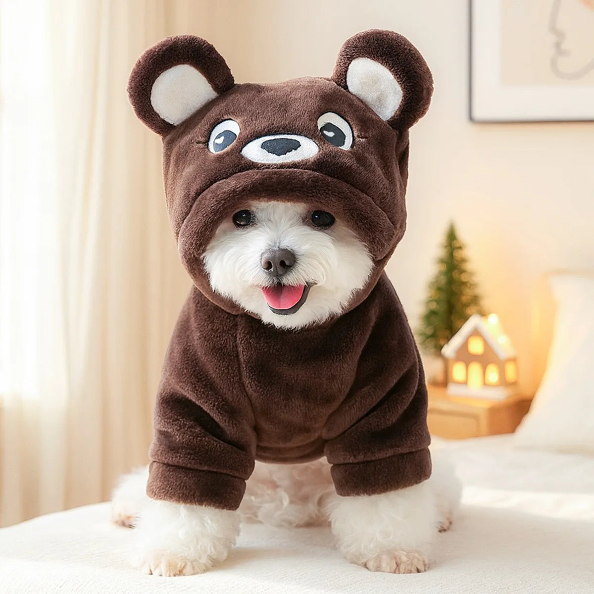 A fluffy white dog wearing a brown bear flannel hoodie sitting on a white surface.