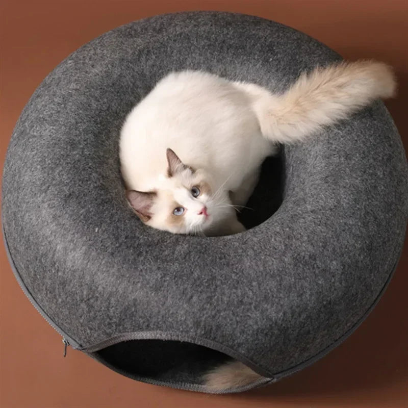 A white and blue cat is peeking out of a round gray felt cat bed with a tunnel hideaway.