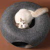 A white and blue cat is peeking out of a round gray felt cat bed with a tunnel hideaway.