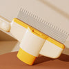 A close-up of a yellow and cream self-cleaning pet brush with massage pins resting on a brown surface.