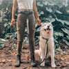 A person wearing brown jeans and boots stands next to a fluffy white dog with a black leash.