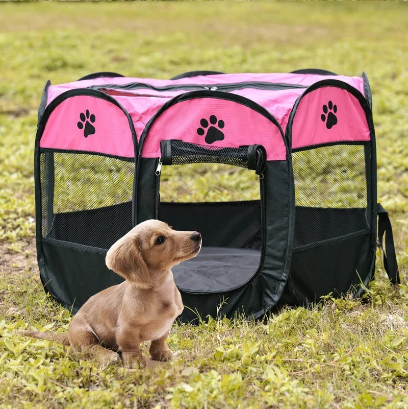 A tan puppy is sitting in a pink and black folding octagonal pet cage outdoors on green grass.