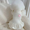 Une robe de princesse blanche pour chien avec bordure duveteuse et détail de cœur rose, présentée sur une surface blanche tex