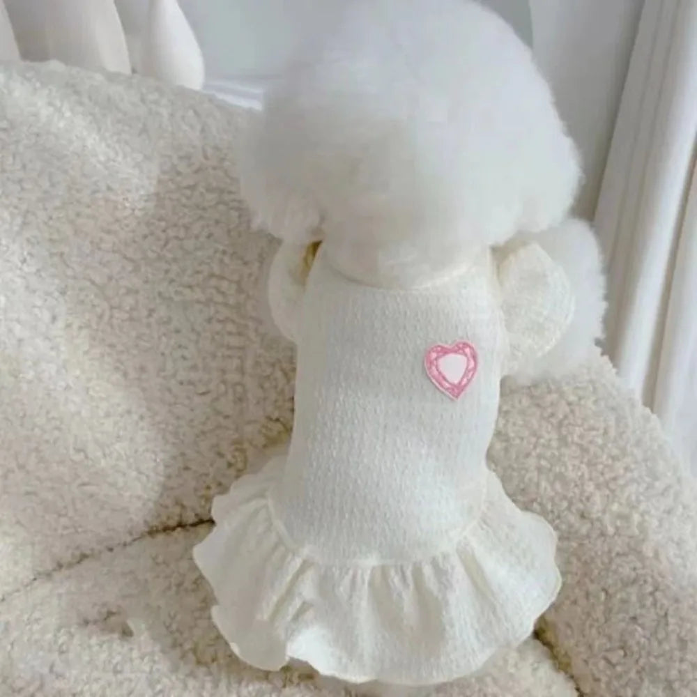 Une robe de princesse blanche pour chien avec bordure duveteuse et détail de cœur rose, présentée sur une surface blanche tex