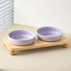 A purple ceramic cat and dog bowl is shown resting on a light wooden stand.