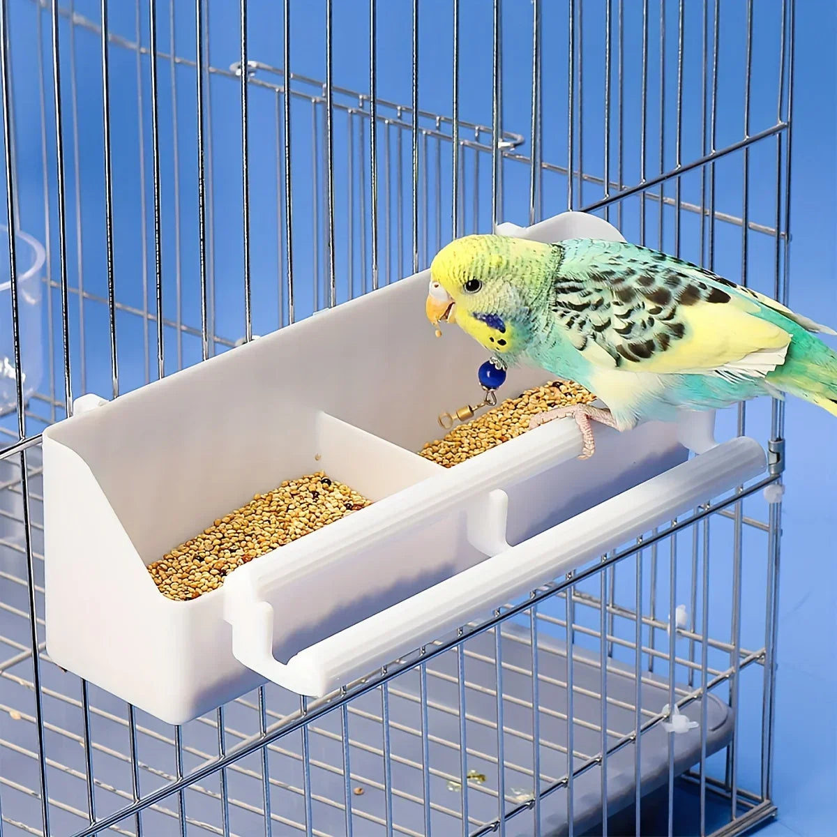 Un perroquet budgerigar jaune et vert nourri d'un distributeur automatique de nourriture blanc dans une cage en fil de fer.