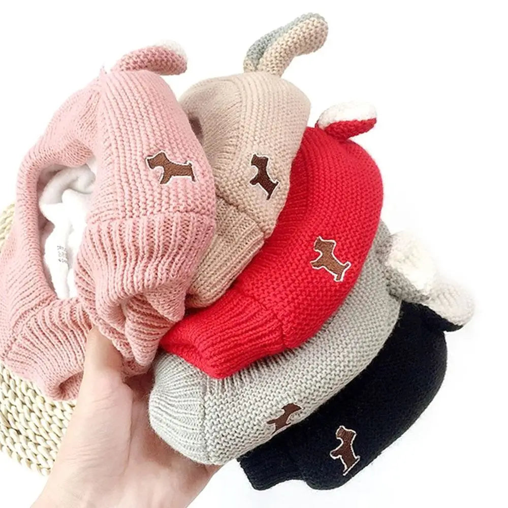 A hand holds a set of cozy knit ear muffs for dogs in pink, beige, and red colors.
