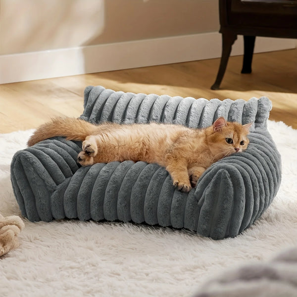 Un chat orange et duveteux se détend sur un lit canapé gris texturé posé sur un tapis blanc.