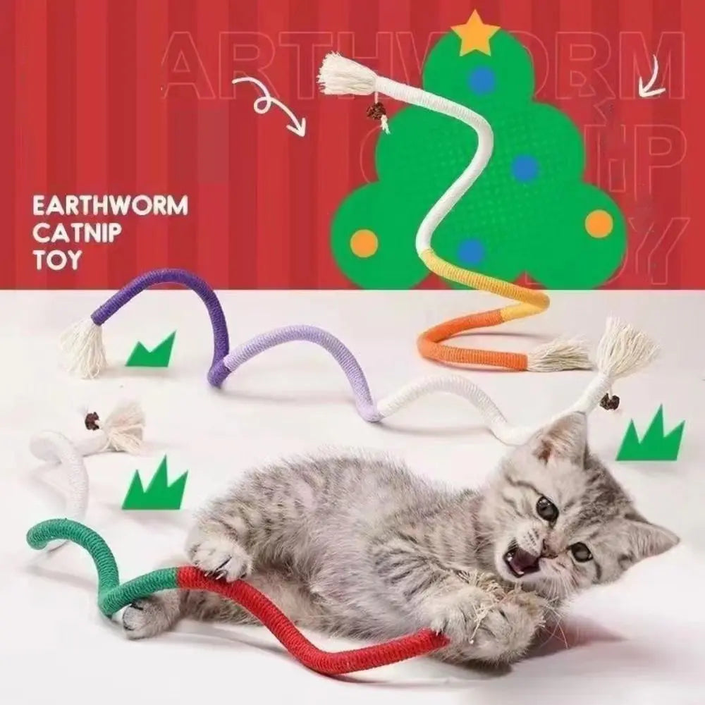 A gray kitten is playing with an earthworm catnip toy made of cotton rope in front of a green Christmas tree background.
