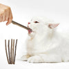 A white cat is licking a stick while another hand holds it, with several natural herbal cat chew sticks nearby.