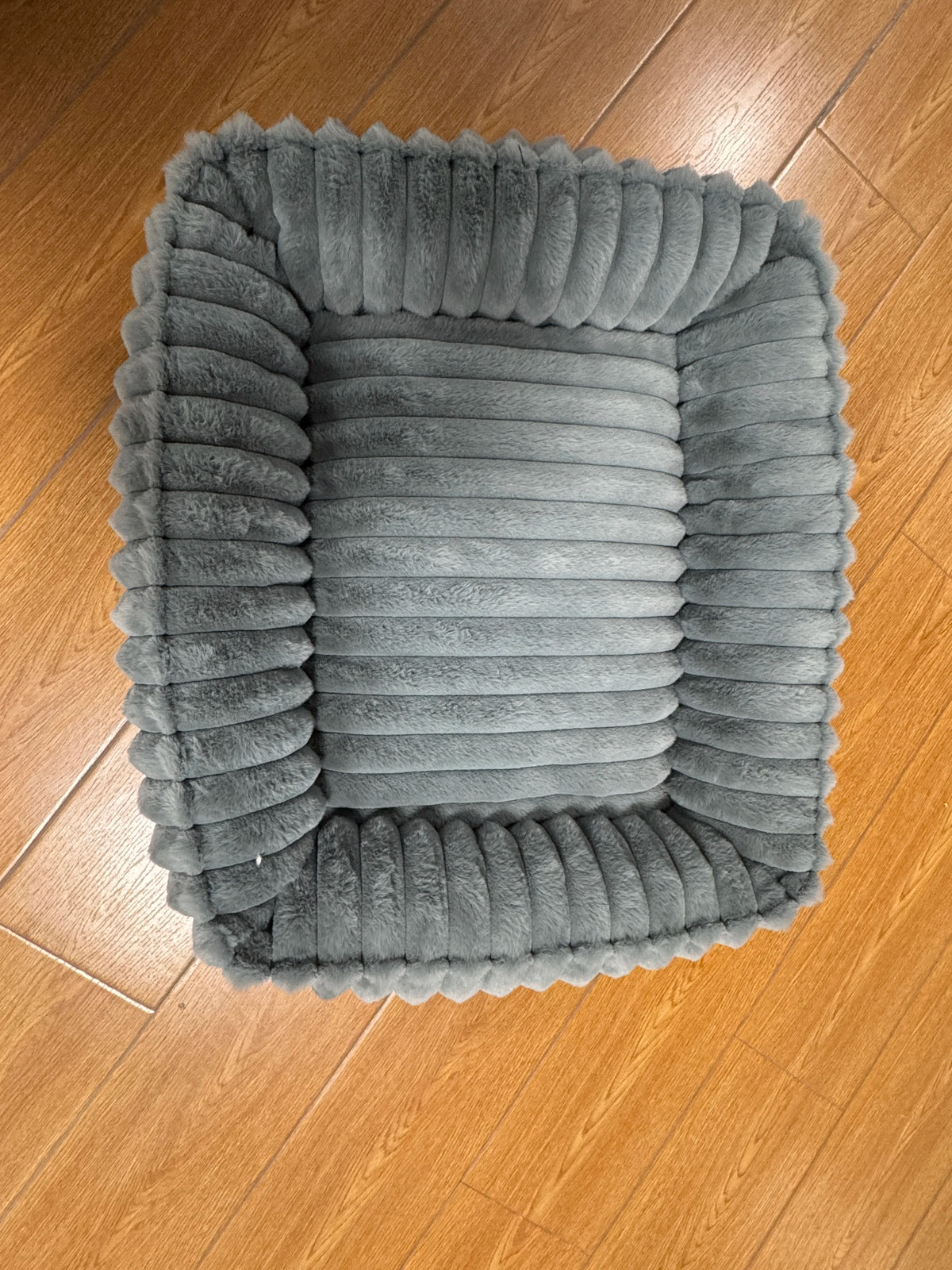 Un lit pour animal de compagnie gris moelleux, carré, avec des côtés à nervures verticales, repose sur un plancher en bois.