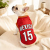 A light-colored dog wearing a red soccer jersey with the word MEXICO and the number 15, lying on a white blanket.