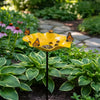 A yellow stainless steel bird feeder filled with seeds is hanging from a stake in a lush garden setting.