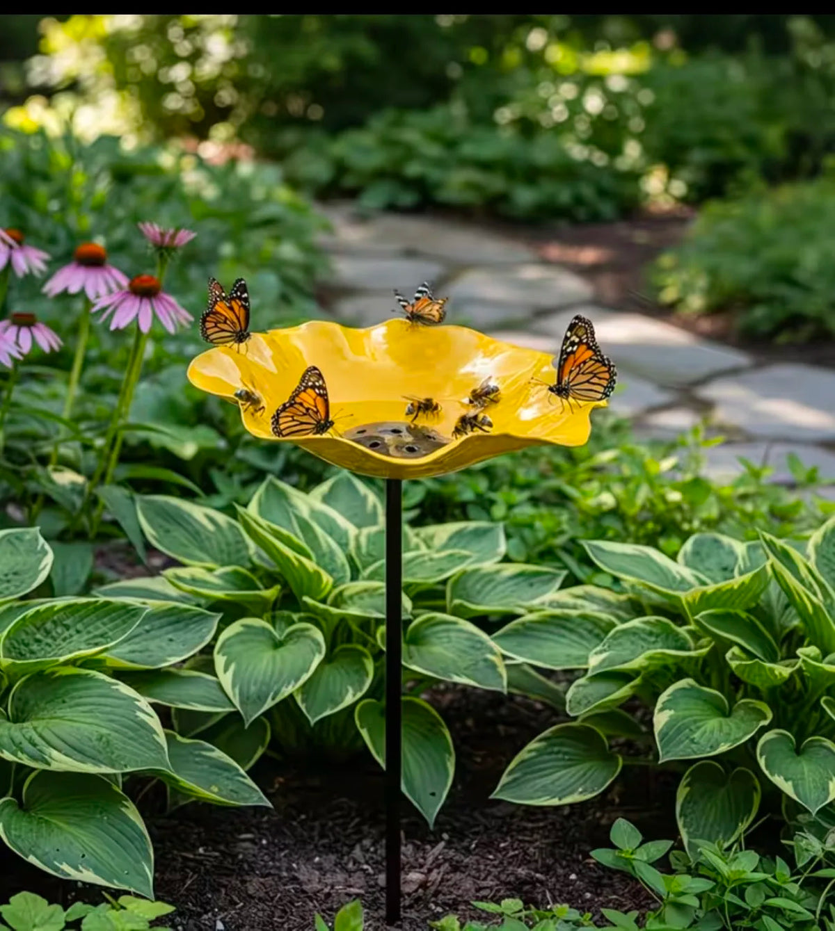 A yellow stainless steel bird feeder filled with seeds is hanging from a stake in a lush garden setting.