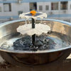 A solar-powered hanging bird bath fountain with a glass bowl, featuring a white, leaf-shaped structure and an orange solar pa