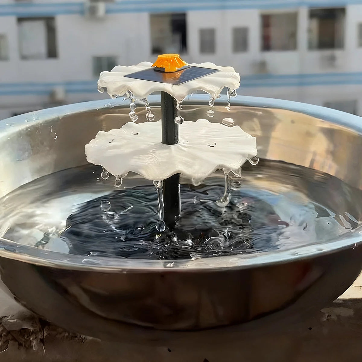 A solar-powered hanging bird bath fountain with a glass bowl, featuring a white, leaf-shaped structure and an orange solar pa
