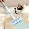 A hand is using a light blue reusable dog hair brush to remove hair from a white cat's fur on a beige surface.