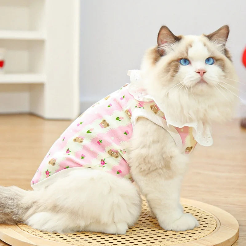 A fluffy white cat wearing a pink cotton vest with a lace collar and cartoon print, sitting on a wooden mat.