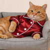 Un gato atigrado naranja vistiendo una camisa de satén roja con lunares blancos y un cuello blanco, sentado en una caja gris.
