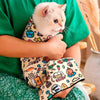 A person is holding a small white cat wrapped in a patterned fabric pet swaddle.